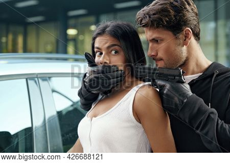 Angry robber closing mouth and threatening with pistol to scared girl. European man and young brunette woman stand near automobile. Male bandit wear hoodie. Concept of robbery and kidnapping