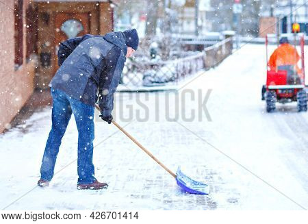 Man With Snow Shovel Cleans Sidewalks In Winter During Snowfall. Winter Time In Europe. Young Man In