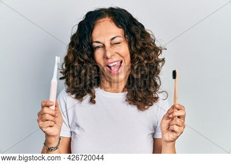 Middle age hispanic woman holding electric toothbrush and teethbrush winking looking at the camera with sexy expression, cheerful and happy face. 