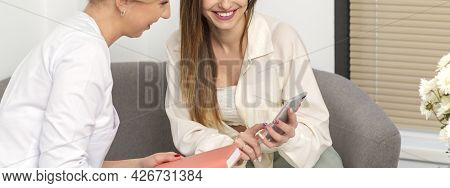 Two Young Women Doctor With Pacient Smiling And Looking At Smartphone Sitting In Hospital Office, Co