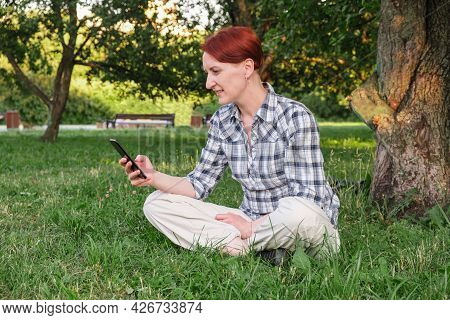 Young Woman With Short Red Hair Sits On The Lawn In The Park And Is Texting On Her Smartphone