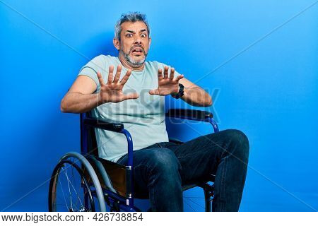Handsome middle age man with grey hair sitting on wheelchair afraid and terrified with fear expression stop gesture with hands, shouting in shock. panic concept. 