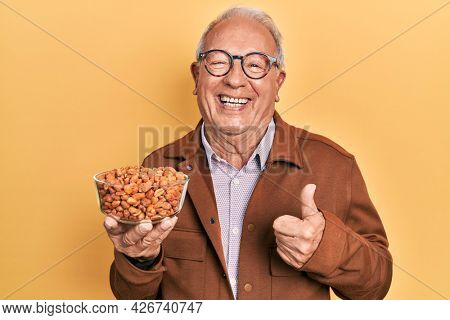Senior man with grey hair holding fried peanuts smiling happy and positive, thumb up doing excellent and approval sign 