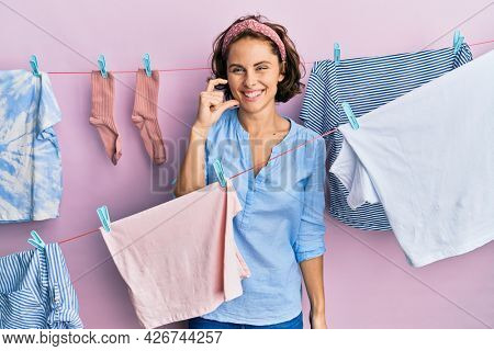 Young brunette woman doing laundry around string hangs smiling and confident gesturing with hand doing small size sign with fingers looking and the camera. measure concept. 