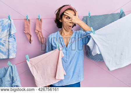 Young brunette woman doing laundry around string hangs peeking in shock covering face and eyes with hand, looking through fingers with embarrassed expression. 
