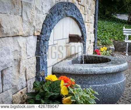 Drinking Fountain Surrounded With Flowers. Stone Basin, Water Running From The Tap. Ladek Zdrój Mine