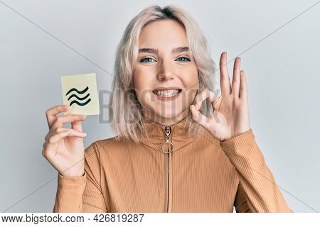 Young blonde girl holding paper with aquarius zodiac sign doing ok sign with fingers, smiling friendly gesturing excellent symbol 