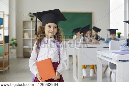 Smiling Schoolgirl In Graduation Cap Holds An Orange Book.