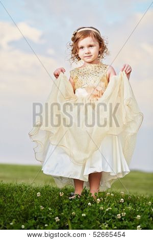 Little girl in beautiful beige gown and crown on head stands  on grassy meadow, lifting up slightly hem of external skirt