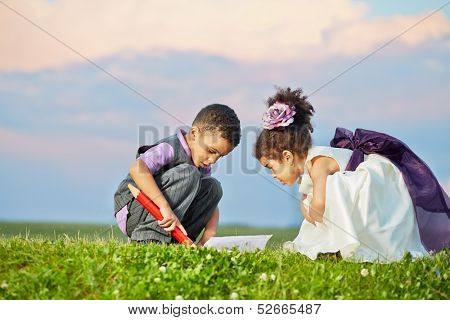 Little boy in grey suit and little girl in light gown with violet waistband sit squatted and examine open copybook that lies on grass
