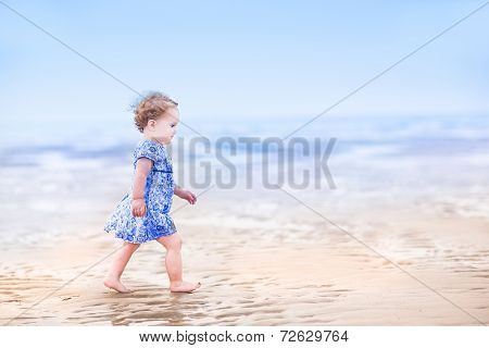Cute Toddler Girl In A Blue Dress Walking On A Beach At Sunset