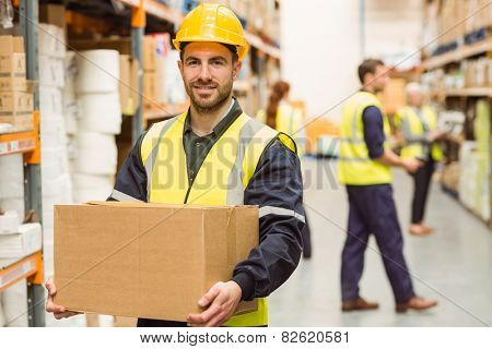 Warehouse worker smiling at camera carrying a box in a large warehouse