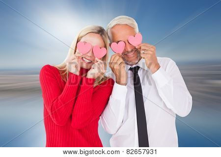 Silly couple holding hearts over their eyes against room with large window looking on city