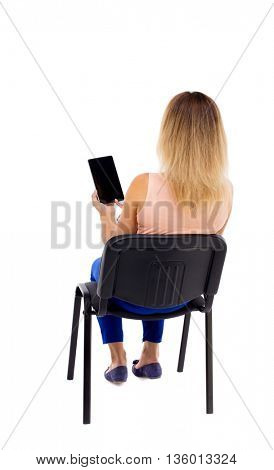 back view of woman sitting on chair and looks at the screen of the tablet.  Rear view people collection.  Blonde in blue pants sitting on an office chair, looking at tablet computer.