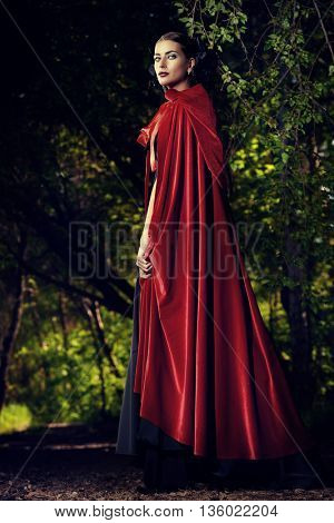 Beautiful brunette woman in black old-fashioned dress and red cloak walking in the thicket of the magic forest. Gothic style. Fashion.