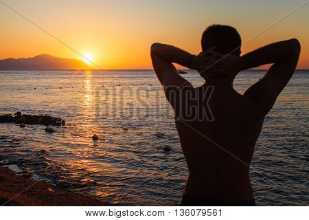 Young man standing with hands behind his head enjoying beautiful colorful sunrise or sunset sea landscape and fresh air