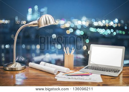 Front view of creative designer desktop with blank white laptop stationery items and table lamp on blurry night city background. Mock up