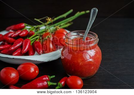 Glass jar of homemade tomato sauce with fresh ingredients on dark wooden background. Natural homemade sauce of tomatoes peppers and vegetables. Chilli jam. Selective focus.