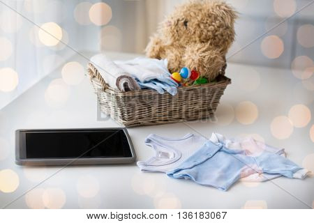 babyhood, motherhood, clothing, technology and object concept - close up of baby clothes and toys for newborn boy in basket with tablet pc computer at home