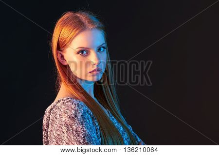 Closeup portrait of beautiful red haired girl lookingat the camera over dark background, in colorful lighting, with copy space