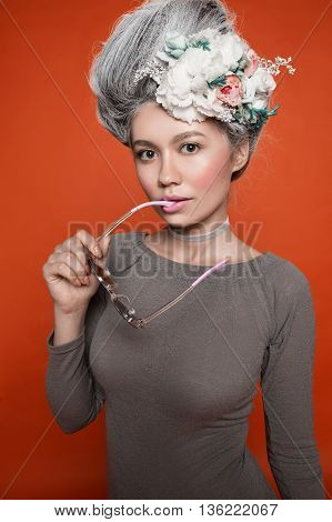 Beautiful girl with nude make up, natural browes, pink lips. White grey hair with diffrent flowers, sunglasses in her mouth