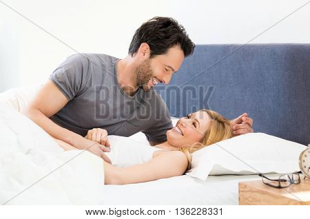 Happy young adult couple smiling in bed. Young couple looking at each other and embracing while lying in bed. Beautiful young couple laughing in bed wearing pajamas.