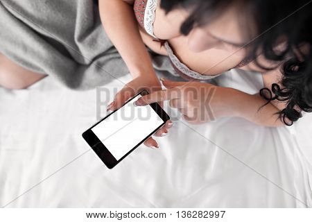 Naked brunette touching cell phone with empty screen. Top view on nude woman in bed using her smartphone. Mobile phone screen with copyspace