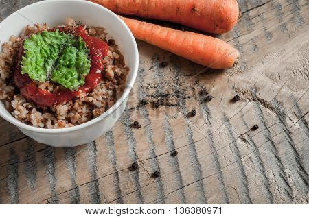 Buckwheat porridge with tomato sauce and leaf salad in a white bowl on a wooden table next to a ceramic bowl with dish is carrots or sliced carrot. Food background. Nice and tasty atmosphere.