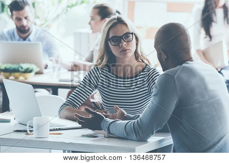 Discussing business. Two young business people discussing something while sitting at the office desk together while their colleagues sitting in the background