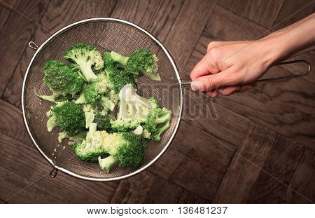 Top view of a hand holding a metal cullender with broccoli against wooden background