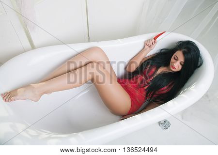 beautiful young adult brunette woman with long hair lying seductively in a white bath smiling and holding a red hot peppers