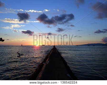 Rock Pier leads to Sunset over the Pacific ocean on the water with birds flying in the foreground and boats off the coast of Oahu Hawaii with Waianae Mountain range visible. February 2016.