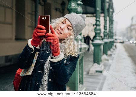 Young blond curly female tourist in warm clothes and red gloves photographs or taking a selfie on city street, winter, snow