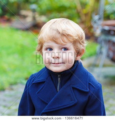Happy little blond toddler boy walking on cold day in blue coat. Funny kid wearing wam clothes on cold autumn or spring day. Carefree childhood. Child having fun outdoors