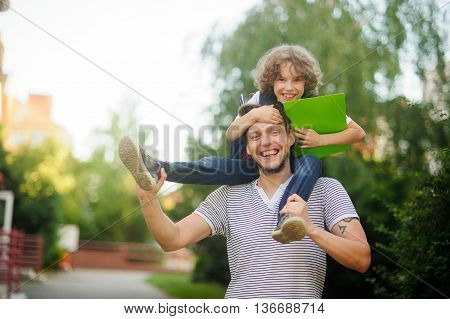 Little schoolboy sitting on the shoulders of his father and clings to his head. The father holds the boy's legs. The boy delighted. He likes to joke with dad. Father and son smiling happily. Back to school