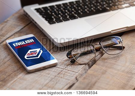 Learning english. Wooden desk with computer, glasses and mobile phone with english dictionary app in the screen.