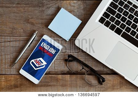Top view of wooden desk, with mobile phone, pen, glasses, post it, and computer. English dictionary application in the phone screen.