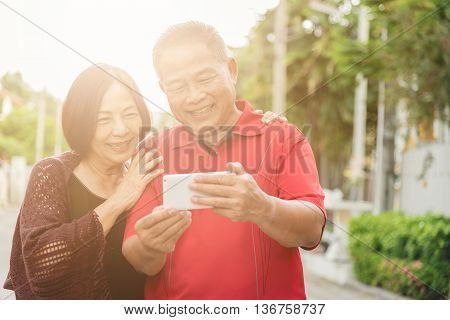 Happy senior Asian couple laughing tigether with mobile smartphone. Warm tone photo with sunlight