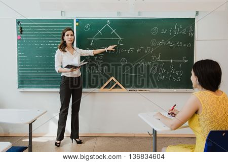 Teacher or docent or educator giving while lesson in front of a blackboard or board a sheet of paper and educate or teaching students or pupils or mates in a school or class.