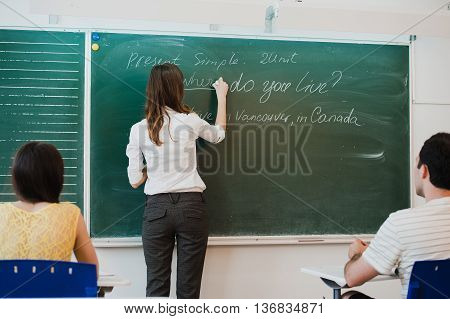 Teacher or docent or educator giving while lesson in front of a blackboard or board a sheet of paper and educate or teaching students or pupils or mates in a school or class.