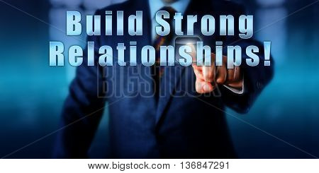 Businessman is touching the phrase Build Strong Relationships! on a virtual monitor. Motivational appeal business concept and call to action. Close up torso shot facing frontally. Blurred background.