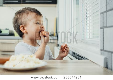 Handsome Boy Shaggy And Shkod Eats Sausage In The Kitchen In A White T-shirt In The Afternoon