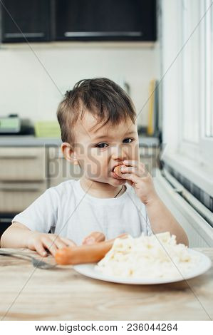 The Child Is Sitting In The Kitchen And Greedily Eats Sausage