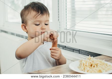 The Child Is Sitting In The Kitchen And Greedily Eats Sausage