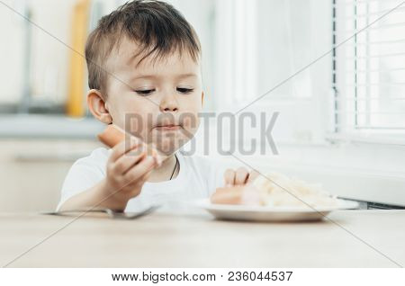 The Child Is Sitting In The Kitchen And Greedily Eats Sausage