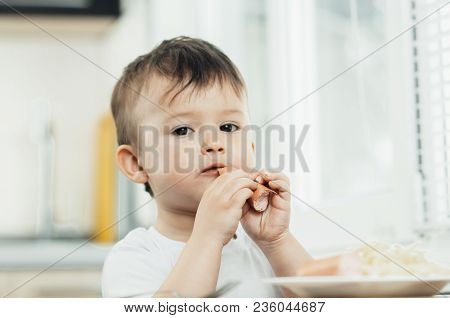 The Child Is Sitting In The Kitchen And Greedily Eats Sausage