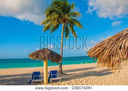 Holguin, Cuba, Playa Esmeralda. Umbrella And Two Lounge Chairs Around Palm Trees. Tropical Beach On 