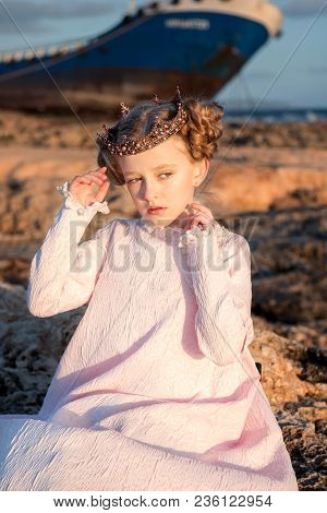 Portrait Of A Fidgety Princess In A Pink Dress And In A Crown That Sits On A Rock Near A Ship Broken