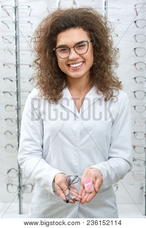 Photo Of Smiling Doctor Helping To Choose Glasses, Standing In Front Of Transparent Stand With Diffe
