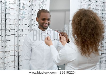 Frontview Of Smiling Man Trying On Eyeglasses In Optical Shop. Young Doctor Is Helping The Client To
